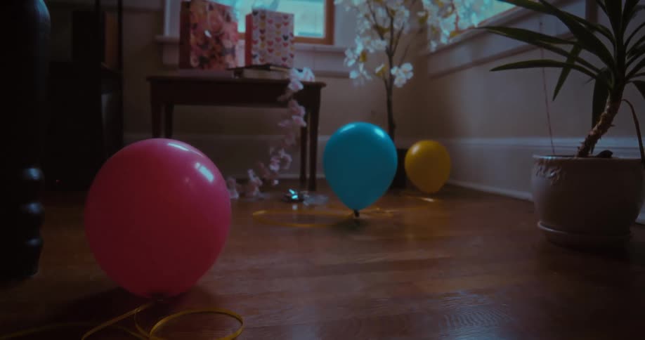 A still life shot of colorful party balloons on a wooden floor in a dimly lit room with gifts in the background