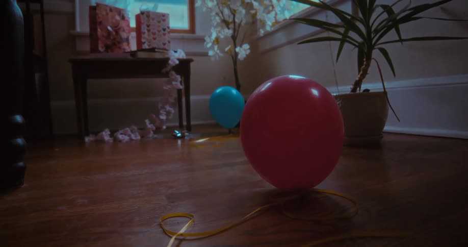 Colorful balloons rest on a hardwood floor in a dimly lit room after a birthday party
