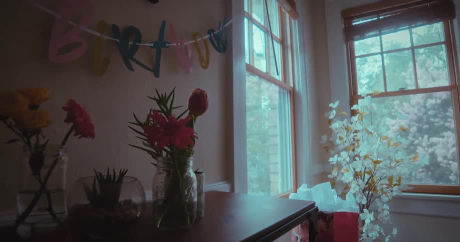 A still life of birthday decorations with flowers, a banner, and a yellow balloon in a room lit by a window
