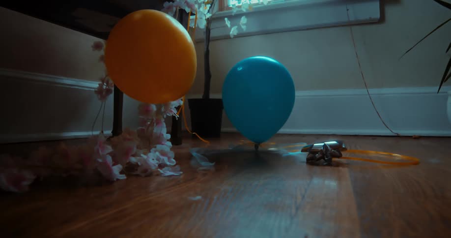 An orange balloon slowly floats down to join a blue balloon on a hardwood floor in a dark room