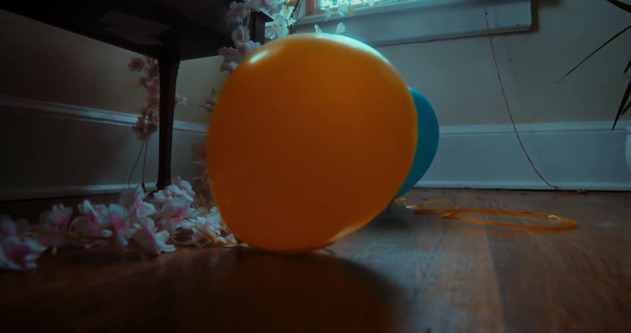 An orange balloon floats down to join a lonely blue balloon on a wooden floor in a dimly lit room after a party
