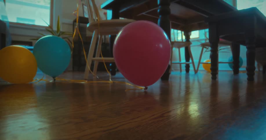 Colorful party balloons gently bob on a polished hardwood floor in a quiet, empty room