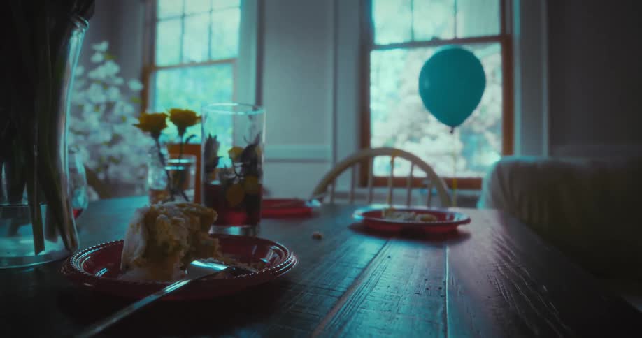 A lone blue balloon floats and then falls over the remains of a birthday party in a dimly lit, empty room