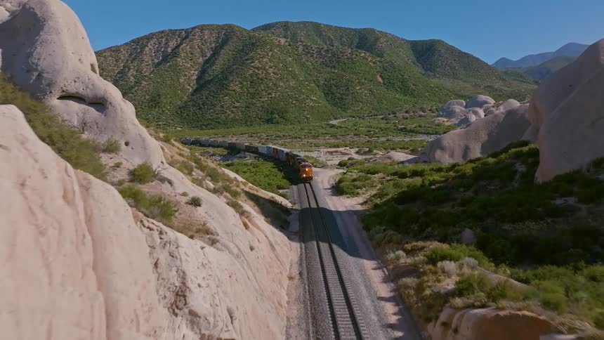An aerial shot flies forward through a rocky canyon to reveal a long freight train traveling through a vast, mountainous landscape on a sunny day