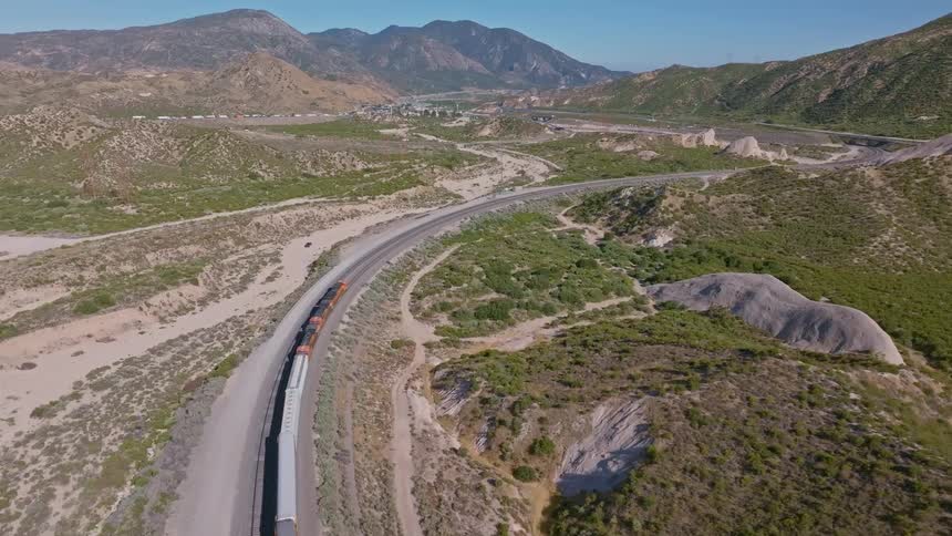 An aerial drone follows a long freight train as it travels on a curving track through a vast, arid valley surrounded by mountains