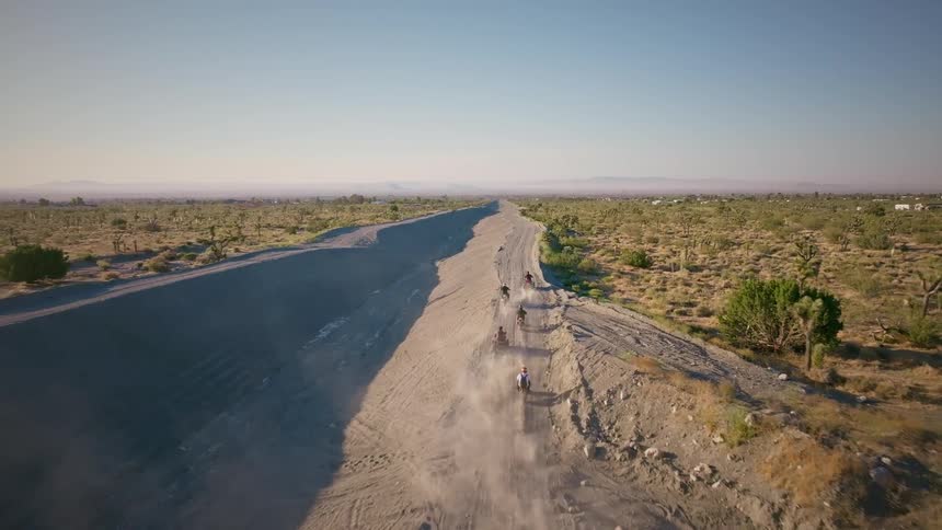 An aerial view shows a group of dirt bikers and ATVs riding along a long, dusty road through the desert landscape