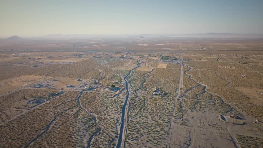 An aerial view starts above a dry flood channel in a desert town, then pulls back to reveal the expansive, arid landscape reaching to distant mountains