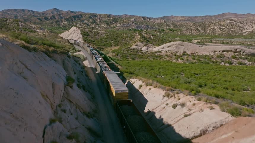 Aerial footage follows a long freight train carrying cargo through a remote and rocky desert canyon on a sunny day