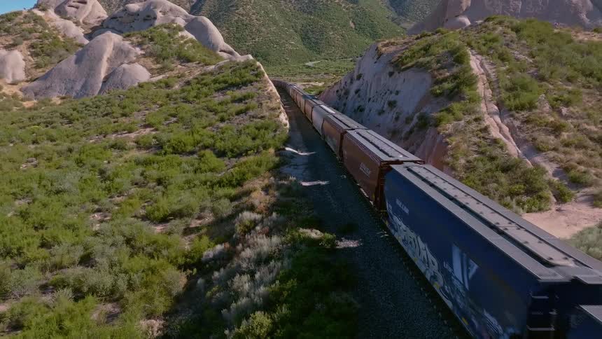 An aerial view captures a long freight train traveling through a remote and rugged mountain landscape with unique rock formations