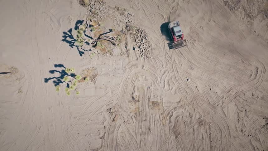 An aerial top-down shot follows a compact track loader as it slowly grades and levels a large patch of sandy desert soil