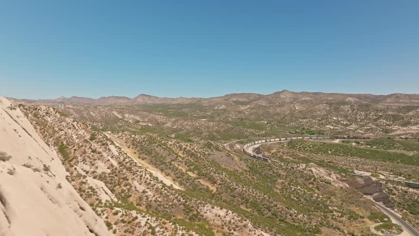 An epic aerial journey over unique sandstone formations reveals a freight train winding through the vast California desert landscape