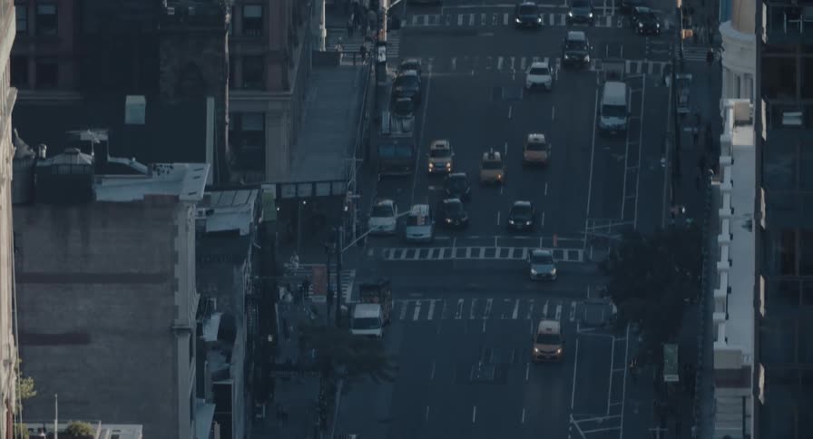 A high-angle long shot tilts up from a city intersection to reveal a bustling New York City avenue with traffic flowing between tall buildings at dusk