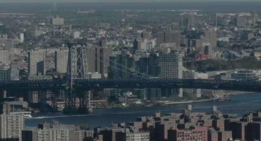 A static telephoto shot of the Williamsburg Bridge with a train crossing and a ferry on the East River on a sunny day
