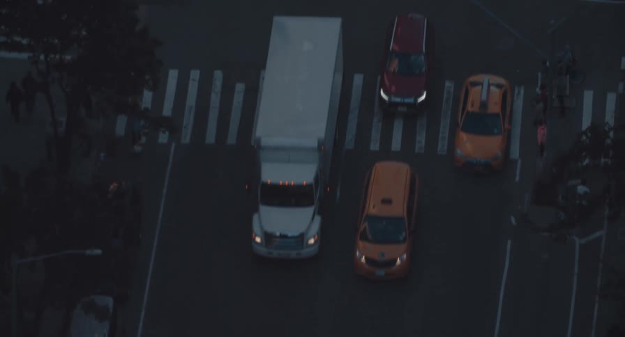 A moody, top-down aerial view tracks the flow of city traffic and pedestrians on a New York street at dusk