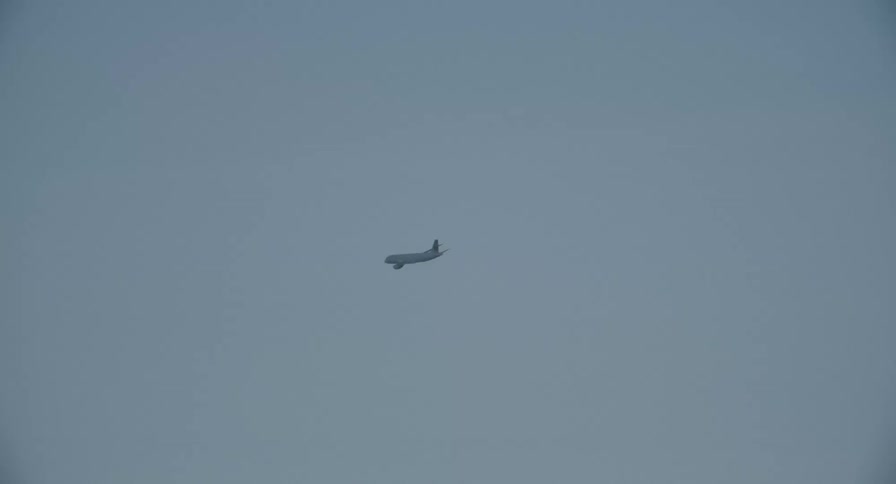 A distant commercial jet flies across a hazy blue sky and exits the frame