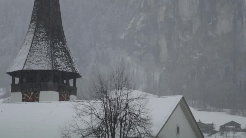 Snow is falling over a building and a clocktower in a mountainous area