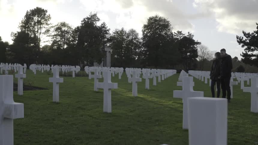 A man and woman stand together looking at a cross in a cemetery