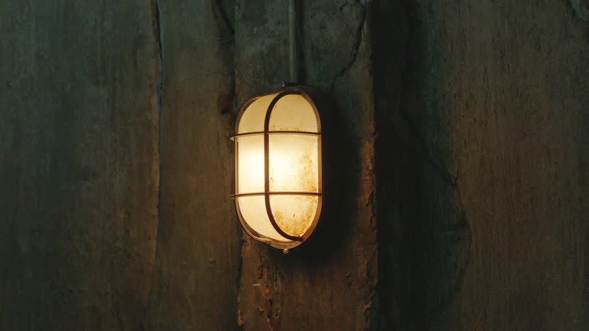 A rusty caged industrial light flickers on a dark, textured concrete wall, casting an eerie and suspenseful glow