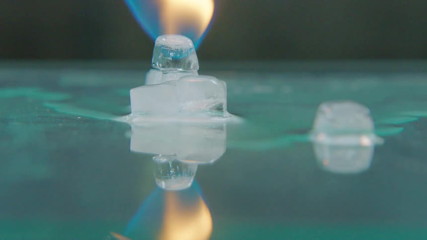 A blowtorch flame melts ice cubes in slow motion, creating a visual contrast between fire and ice on a reflective surface