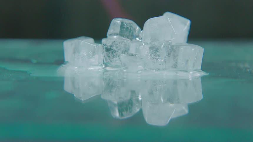 Timelapse of stacked ice cubes melting on a reflective teal surface with a heat torch accelerating the process