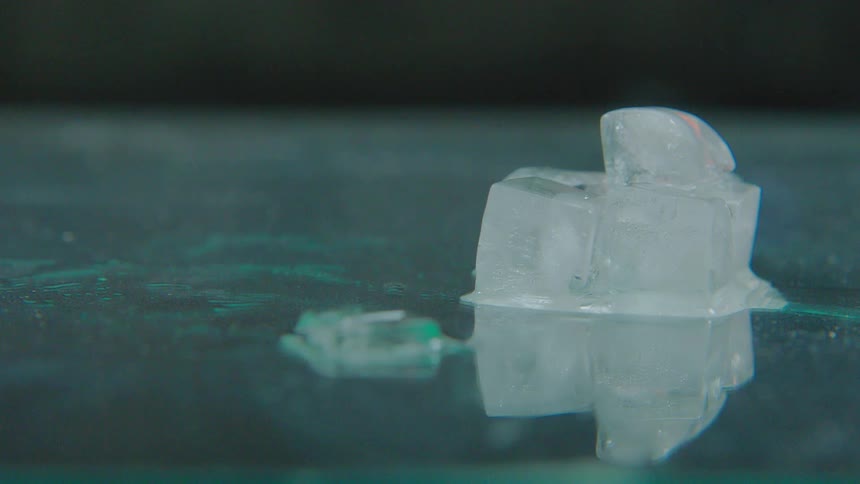 A macro timelapse of a stack of ice cubes melting into a puddle of water on a reflective surface