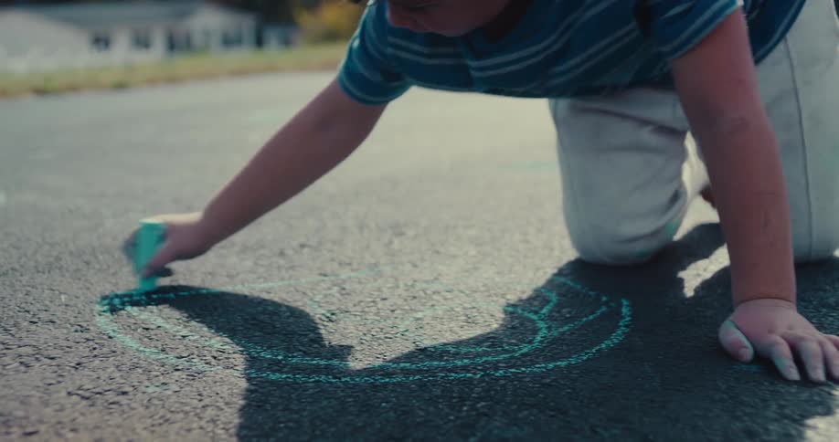 A barefoot boy draws with sidewalk chalk on a driveway on a sunny afternoon in slow motion