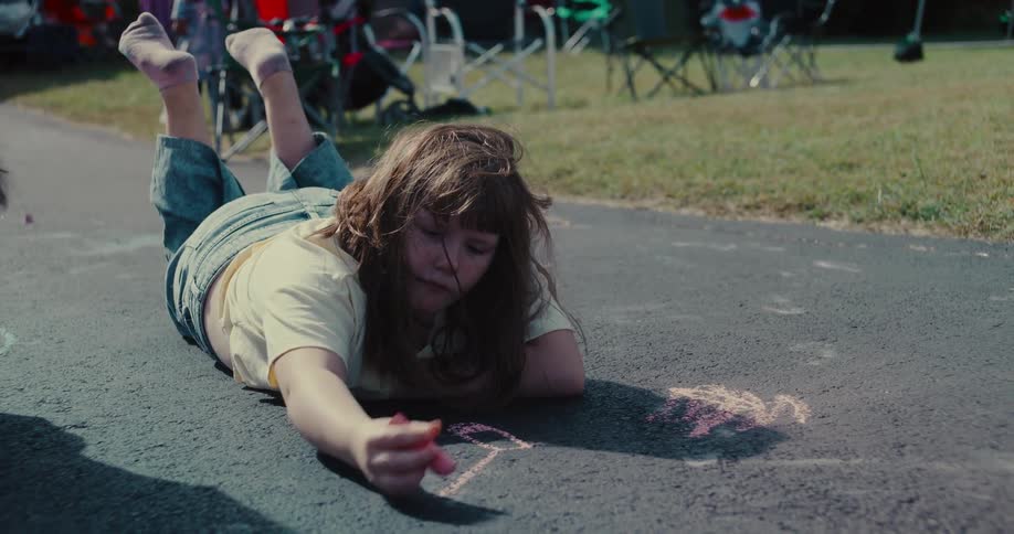 A young girl lies on a paved driveway and draws with pink sidewalk chalk on a sunny day