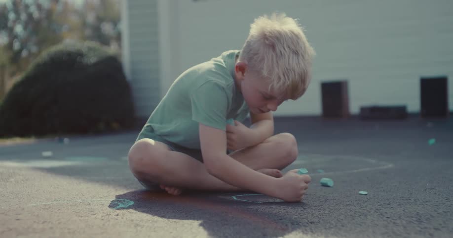 A young blonde boy sits cross-legged on an asphalt driveway, intently drawing with a piece of blue chalk