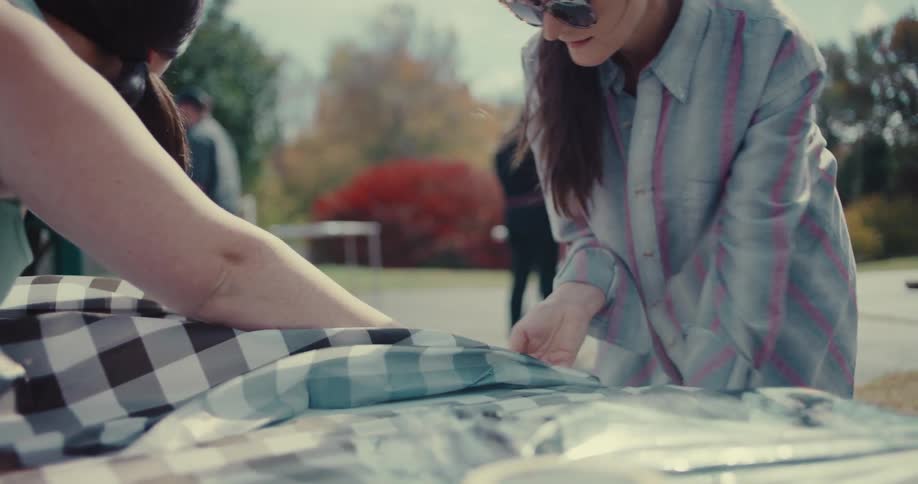 Two women work together to lay out a black and white checkered picnic tablecloth in slow motion