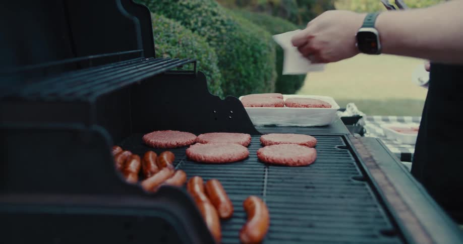 A person carefully places a raw hamburger patty on a hot grill next to sizzling sausages during a backyard barbecue