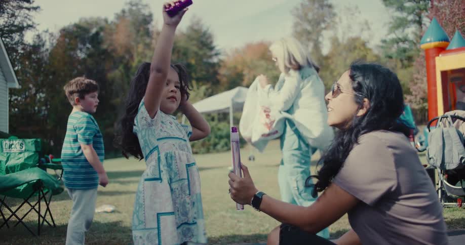 In slow motion, a mother helps her young daughter create bubbles with a large bubble wand at a sunny outdoor party