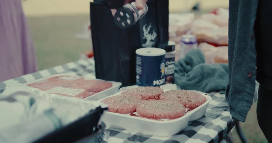 Close-up slow-motion shot of seasoning raw hamburger patties on a checkered tablecloth for an outdoor barbecue
