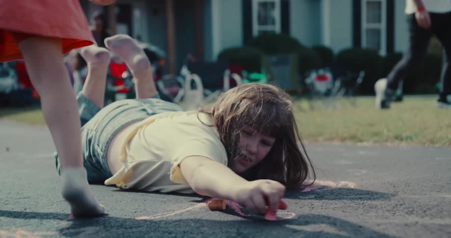 A young girl lies on a driveway, focused on drawing with pink sidewalk chalk on a sunny day in slow motion
