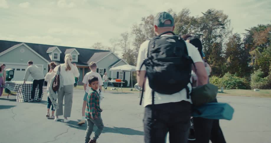 A diverse group of families and friends walk towards an outdoor community festival on a bright sunny day