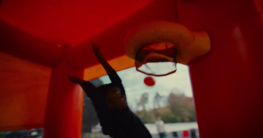 A young boy plays basketball in slow motion inside a red and yellow bouncy house in a suburban backyard