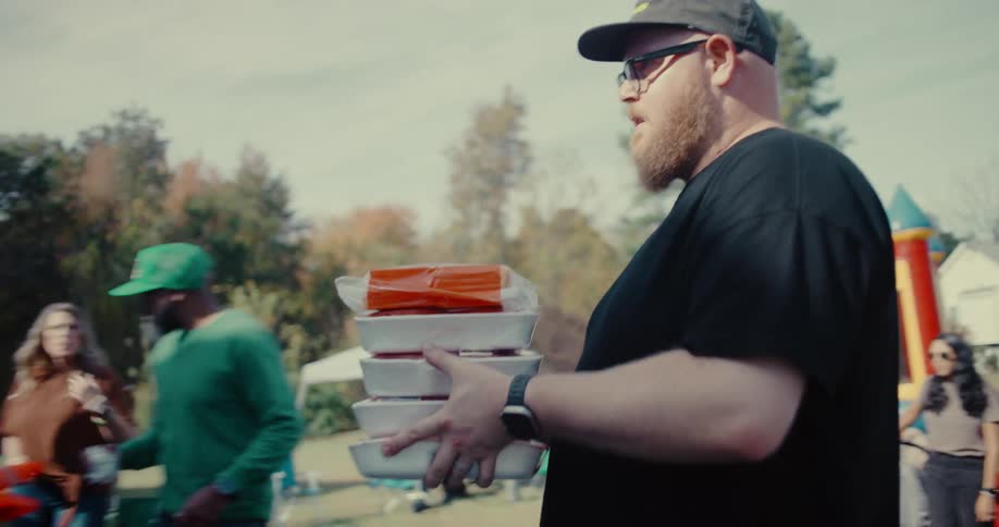 A bearded man carries a stack of uncooked burgers and hot dogs to the grill area during a sunny backyard party in slow motion