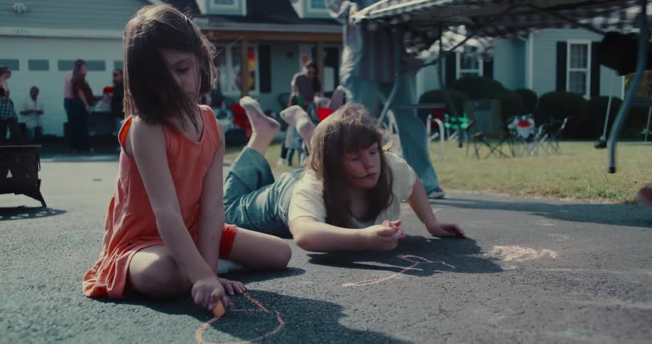 Two young girls draw with colorful chalk on the driveway of a suburban home during a neighborhood party