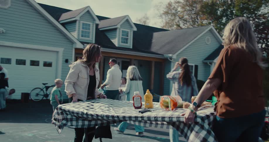 Two women place a checkered tablecloth on a table as neighbors gather for a festive block party in a suburban driveway