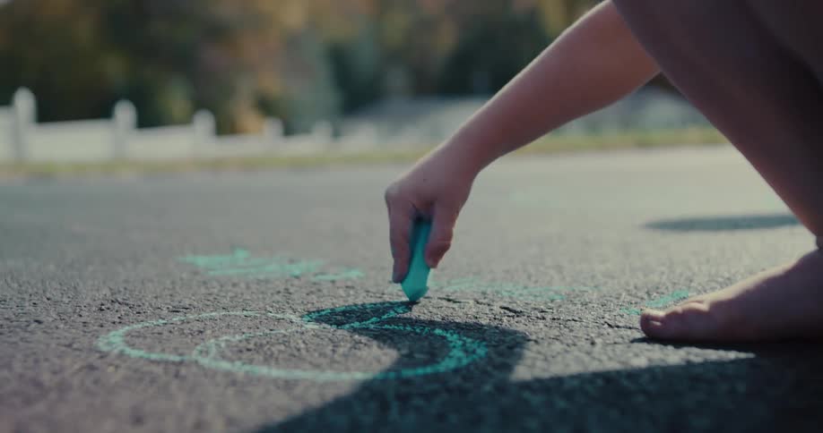 A barefoot child draws on the asphalt with turquoise chalk in slow motion on a sunny day