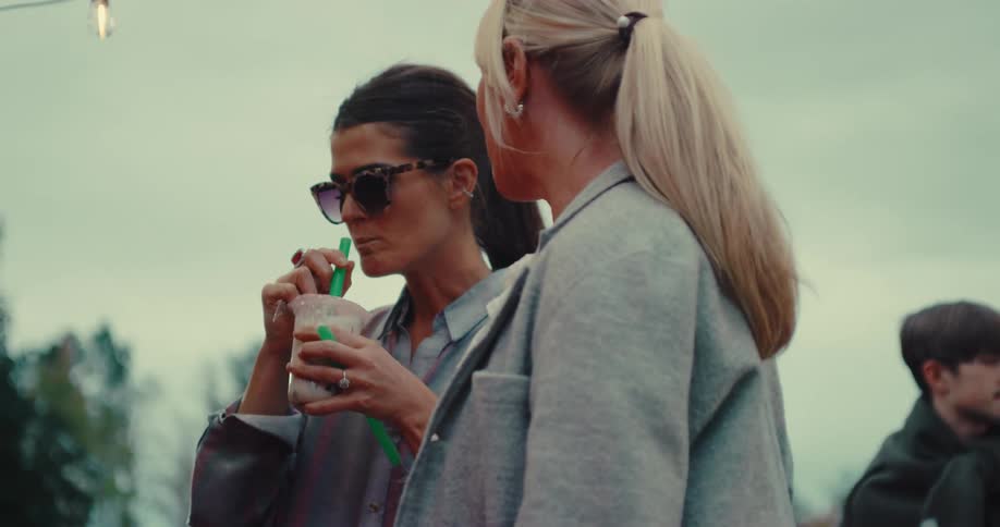 Two stylish women share a bubble tea at an outdoor gathering in slow motion