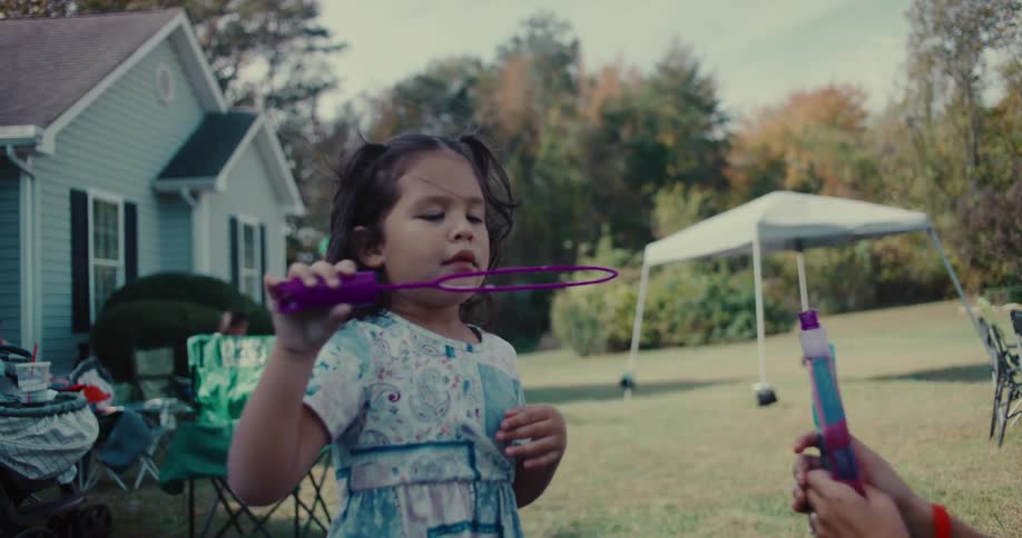 A young girl in a pretty dress concentrates on blowing bubbles in her backyard with her mother in slow motion