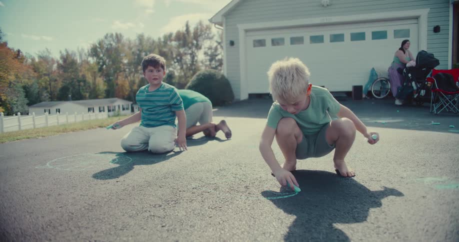 Three young boys spend a sunny afternoon drawing with colorful chalk on their suburban driveway while their family watches nearby