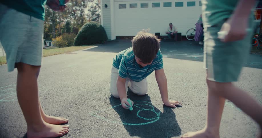 Slow-motion shot of a boy kneeling on the asphalt to draw with chalk as his brothers play nearby