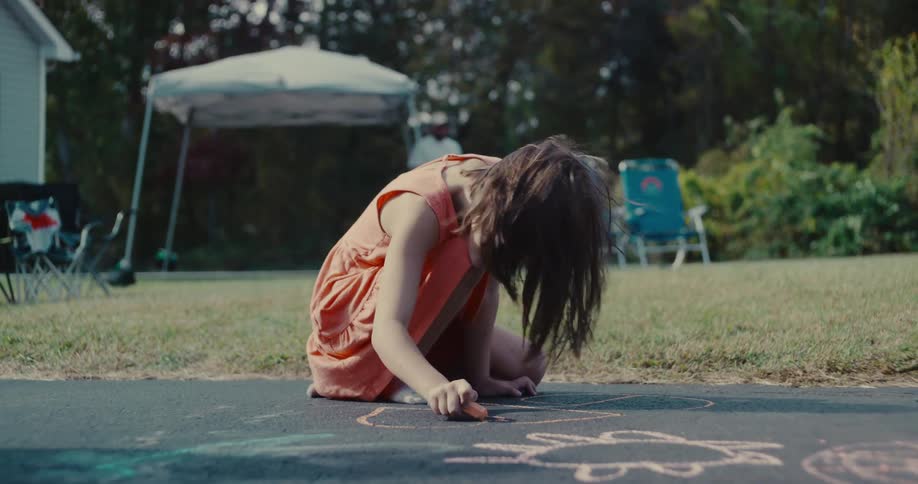 Slow motion shot of a young girl drawing with chalk on the driveway before getting up and running to play at a backyard party