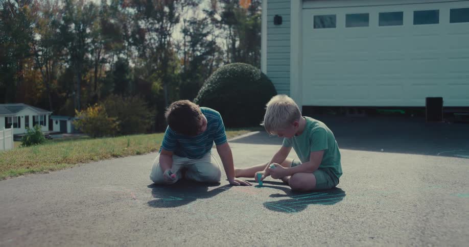 Two young brothers have fun drawing with sidewalk chalk in their driveway, with one boy playfully drawing on his leg before jumping up to run around