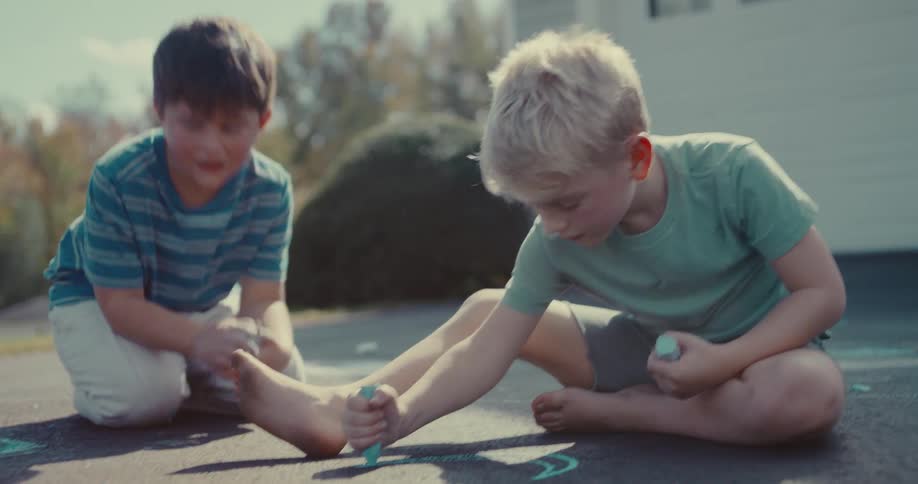 Barefoot and focused, a young boy creates chalk art on the pavement with his friends on a summer day