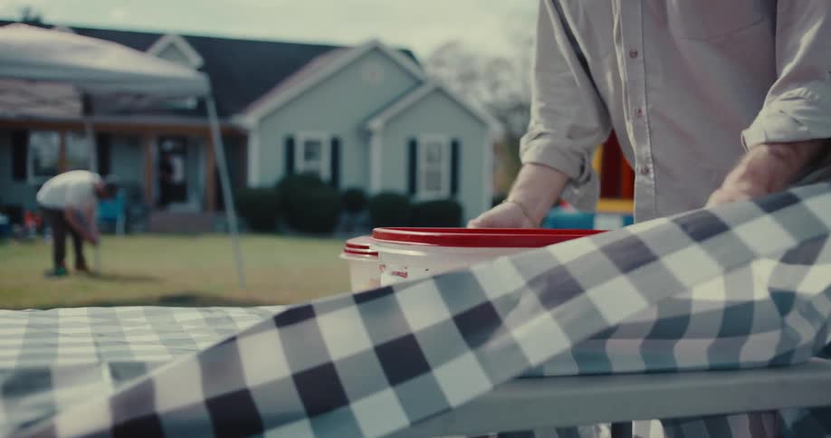 A person sets down containers of cupcakes in slow motion on a checkered tablecloth while preparing for a backyard party