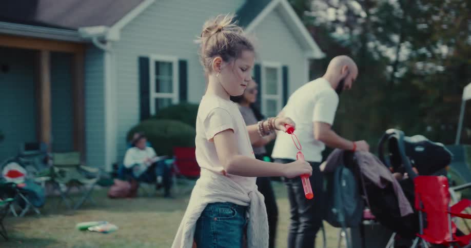 A young girl with her hair in a bun creates large soap bubbles with a bubble wand in slow motion during a backyard party