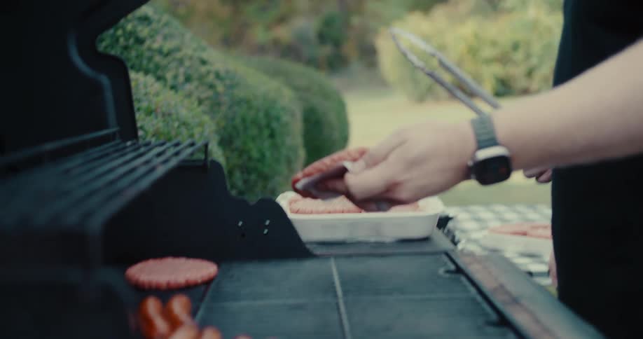 Slow motion shot of a man placing seasoned raw burger patties on a hot backyard barbecue grill
