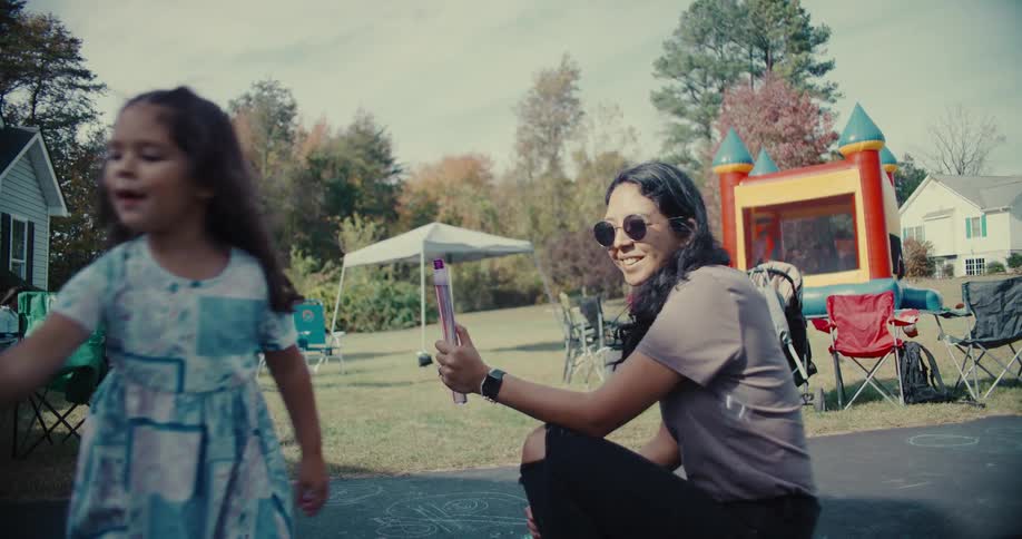 A happy mother and young daughter play with a giant bubble wand in slow motion at a sunny backyard party
